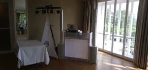 Ein kleines Indoor-DJ-Setup für eine Hochzeit steht neben großen Fenstern mit Vorhängen. Es umfasst Lautsprecher, einen Laptop auf einem Ständer und eine dreieckige Flagge mit einem Logo. Links steht ein mit einem weißen Tuch bedeckter Tisch. Sonnenlicht fällt hinein, beleuchtet den Holzboden und schafft das perfekte Ambiente für eine Hochzeit.