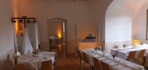 Ein gut beleuchteter Speisesaal mit elegant gedeckten Tischen, weißen Tischdecken und Holzstühlen bietet die perfekte Kulisse für eine Hochzeit. Sanftes gelbes Licht schafft eine warme Atmosphäre, während ein DJ-Pult in der Ecke auf festliche Hochzeitsfeiern hindeutet.