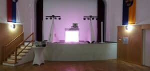 Eine schwach beleuchtete Bühne mit einer hellen Leinwand und Lautsprechern, flankiert von zwei Lichtbannern, schafft die Kulisse für eine Hochzeit. Der Holzboden des Saals glänzt unter den Fahnen an den Wänden. Links steht ein kleiner Tisch mit einer weißen Decke, auf dem Gläser und eine Flasche stehen, perfekt zum Anstoßen während dieser besonderen Feier.