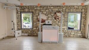 Ein gemütliches DJ-Setup für den Innenbereich vor einer Steinmauer besteht aus zwei dekorativen Kreisen, perfekt für eine Hochzeit. Zur Ausstattung gehören ein DJ-Pult, Lautsprecher und Lichter, und die Fenster bieten Ausblicke auf die üppige Natur. Pfirsichfarbene Blumen verleihen dieser hochzeitsreifen Szene eine romantische Note.