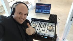 Eine Person mit Kopfhörern streckt vor der DJ-Ausrüstung den Daumen nach oben, perfekt, um bei jeder Hochzeit für Stimmung zu sorgen. Ein Laptop mit Musiksoftware und ein Mischpult stehen auf einem hell gefliesten Boden bereit, umgeben von Stühlen, die auf Gäste warten.