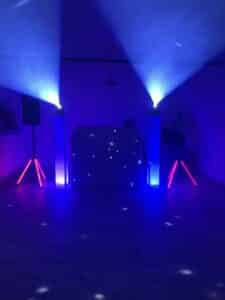 Ein schwach beleuchteter Raum, perfekt für eine Hochzeit, mit einem DJ-Pult, das in blaues und rosa Licht getaucht ist. Zwei Strahlen strahlen nach oben und werfen einen Sterneneffekt auf das Pult. Das Setup umfasst zwei Lautsprecher auf Ständern, die das minimalistische, farbenfrohe Ambiente verstärken, das ideal für Hochzeitsfeiern ist.