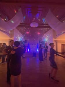 Der schwach beleuchtete Ballsaal strahlt eine romantische Atmosphäre aus, die perfekt für eine Hochzeit ist, mit Paaren, die unter bunten Lichtern im Disco-Stil tanzen. Die hohe Decke ist mit drapiertem Stoff geschmückt und ein DJ-Setup verstärkt die festliche Atmosphäre, ideal für diejenigen, die schon weit mit der Hochzeitsplanung fortgeschritten sind.