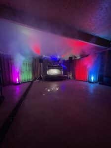 Eine schwach beleuchtete Tanzfläche mit bunten Lichtern und Nebel schafft die Kulisse für eine unvergessliche Hochzeit. Ein DJ-Pult in der Mitte, beleuchtet von roten, grünen und blauen Scheinwerfern, verspricht eine lebendige Partyatmosphäre, die perfekt ist, um die Liebe zu feiern.