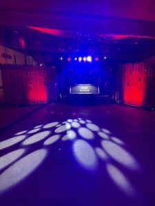 Ein schwach beleuchteter Nachtclub, der sich perfekt für eine einzigartige Hochzeitsfeier eignet, verfügt über ein DJ-Pult mit einem Red Bull-Logo. Der Raum ist mit violetten und roten Lichtern beleuchtet, die komplizierte Muster auf den Boden werfen und eine lebendige und energiegeladene Atmosphäre schaffen, ideal für diejenigen, die die ganze Nacht durchtanzen möchten.