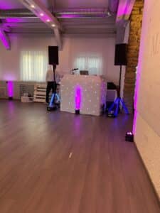 Ein dekorierter Veranstaltungsraum, der perfekt für eine Hochzeit ist, mit einem DJ-Pult, das mit weißem Stoff behangen ist und von kleinen Lichtern funkelt. Der Raum, der sich ideal zum Heiraten eignet, verfügt über Hartholzböden und Stein- und weiße Wände, die von violettem Ambientelicht angestrahlt werden. Die Audioausrüstung steht bereit, und hinter dem Pult ist teilweise jemand zu sehen.