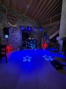 In einem schwach beleuchteten Raum mit Steinwänden, der sich perfekt für eine einzigartige Hochzeit eignet, erstrahlt ein DJ-Pult in blauem Licht. Dekorative Projektionen werfen florale Muster auf den Boden, während Holztreppen und ein Wappen eine zauberhafte Kulisse für Ihre Hochzeitsfeier bieten.