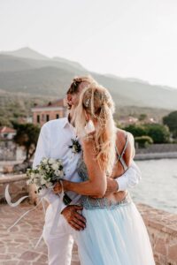 Ein Paar umarmt und küsst sich in der Nähe einer Uferpromenade. Die Frau im hellblauen Kleid hält einen Strauß weißer Blumen und fängt die Essenz der Romantik vor einer Kulisse ein, die perfekt für Hochzeitserinnerungen ist, mit malerischen Bergen und bezaubernden Gebäuden unter einem klaren Himmel.