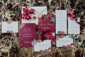 Eine flache Darstellung einer Hochzeitseinladungsreihe auf einer Marmoroberfläche mit Blumenmustern und burgunderfarbenen Akzenten. Enthält Umschläge, Einladungskarten, Ringe und rote Blumen. Ein Wachssiegel und eine Perlenkette verleihen diesem exquisiten Hochzeitsplanungs-Ensemble elegante Akzente.