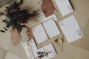 Auf einem gefliesten Boden wird eine Sammlung eleganter Hochzeitspapeterie ausgestellt, die sich perfekt für Ihre Hochzeitsplanung eignet. Sie umfasst mit Blumen bedruckte Karten, Umschläge, eine „Save the Date“-Karte und getrocknete Blumen. In einem braun-weißen Farbschema mit goldenen Akzenten liegen zwei Ringe neben einer offenen Karte.