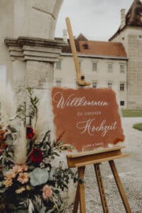 Ein Willkommensschild für eine Hochzeit steht auf einer Staffelei, umgeben von bunten Blumen und Pampasgras. Darauf steht „Willkommen zu unserer Hochzeit“ und darunter „Lara & Daniel“. Ein historisches Gebäude bietet eine bezaubernde Kulisse für diese wunderschön geplante Feier.