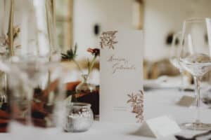 Ein wunderschön gedeckter Tisch mit einer weißen Menükarte mit Blumenmuster, umgeben von elegantem Glasgeschirr und kleinen Blumenarrangements – perfekt für eine Traumhochzeit. Das Datum „25. Juni 2022“ und die Namen „Lara & Daniel“ sind elegant auf das Menü gedruckt und verkörpern eine durchdachte Hochzeitsplanung.