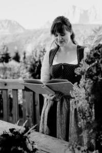 Eine Frau in traditioneller Kleidung steht auf einem Holzdeck und denkt vielleicht über Hochzeitsplanung nach, während sie in einem Buch liest. Im Hintergrund sind eine malerische Berglandschaft und üppiges Grün zu sehen. Ein Blumenstrauß schmückt den Tisch vor ihr. Das Foto ist schwarzweiß.