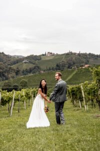 Ein Paar in Hochzeitskleidung hält Händchen und lächelt, während es in einem Weinberg steht. Während ihrer Hochzeit sind die Braut in ihrem weißen Kleid und der Bräutigam in seinem grauen Anzug von üppigen grünen Weinreben umgeben, mit sanften Hügeln als atemberaubender Kulisse.