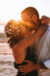 Ein Paar küsst sich bei Sonnenuntergang zärtlich am Strand und feiert seine Hochzeit. Die Frau mit gemustertem Kopftuch und Sonnenbrille und der Mann mit Brille und weißem Hemd genießen die warme, leuchtende Umarmung der Sonne und träumen von ihrem perfekten Tag.