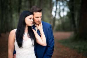 Während ihrer Hochzeit legt eine Frau in einem weißen Spitzenkleid sanft ihre Hand auf das Gesicht eines Mannes, während sie zusammen in einem Wald stehen. Der Mann, in einen blauen Anzug gekleidet, blickt nachdenklich zwischen den hoch aufragenden Bäumen und dem unscharfen Hintergrund hindurch und fängt einen ruhigen Moment der Liebe ein.