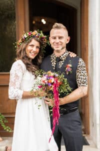 Ein lächelndes Paar steht draußen, bereit zu heiraten. Die Frau trägt ein weißes Spitzenkleid und einen Blumenkranz und hält einen bunten Blumenstrauß. Der Mann trägt ein Hemd mit Leopardenmuster, eine schwarze Weste und eine Krawatte. Sie stehen vor einer Holztür und halten diesen freudigen Moment ihrer Hochzeit fest.