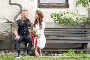 Ein Paar sitzt auf einer Holzbank und lächelt sich an, was die Freude ihrer Hochzeit ausdrückt. Der Mann trägt ein gemustertes Hemd und eine Weste, während die Frau in einem weißen Kleid mit Blumenkrone glänzt und einen Blumenstrauß hält. Eine rustikale Mauer und ein kleiner Baum rahmen diese bezaubernde Szene des Hochzeitsglücks ein.