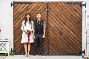Ein Paar steht vor großen Holztüren. Die Frau trägt ein weißes Kleid mit Blumenkrone und Blumenstrauß, während der Mann ein gemustertes Hemd und eine Weste trägt. Ihr Hochzeitsmoment ist wunderschön eingefangen, als sie beide auf einer Betonfläche stehend in die Kamera blicken.