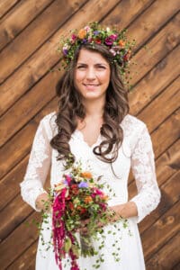 Eine Braut mit langen braunen Haaren steht vor einem hölzernen Hintergrund und verkörpert die Essenz einer Hochzeit. Sie trägt ein weißes Spitzenkleid und eine bunte Blumenkrone und hält einen farbenfrohen Blumenstrauß und Grünpflanzen in der Hand, was perfekte Hochzeitsplanung zur Schau stellt.