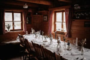 Ein gemütlicher Speisesaal mit Holzwänden und zwei Fenstern, perfekt für eine Hochzeit. Ein langer Tisch ist mit weißen Tischdecken, Glasvasen, Kerzen und zarten Blumenarrangements gedeckt. Rot-weiß karierte Vorhänge verleihen dem warmen Ambiente eine rustikale Note, ideal für eine schöne Hochzeitsplanung.