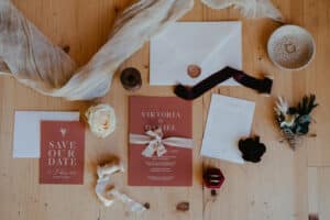 Flache Anordnung von Hochzeitspapeterie auf einer Holzoberfläche, mit einer „Save Our Date“-Karte, einer Einladung mit Schleife, Umschlägen, Wachssiegel, weißen und schwarzen Blumen, Stoff, Ringschachtel und kleiner Schale – perfekte Elemente für jede Hochzeit.
