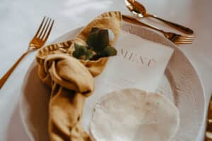 Eine elegante Hochzeitstafel besteht aus einem weißen Teller, goldenem Besteck und einer senffarbenen Serviette. Auf dem Teller liegt ein Menü mit einem grünen Zweig. Das Ambiente lässt auf ein gehobenes Speiseerlebnis schließen, perfekt für die Hochzeitsplanung.