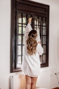 Eine Frau im weißen Morgenmantel steht auf Zehenspitzen und hängt ein Brautkleid ans Fenster – ein heiterer Moment inmitten der Hochzeitsplanung. Ihr langes Haar ist zu Zöpfen geflochten. Der Raum ist sanft beleuchtet, mit einem Fenster mit Holzrahmen und einem Heizkörper darunter.