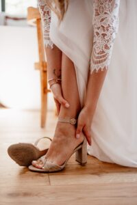 Eine Person in einem weißen Spitzenkleid, die sich vielleicht auf ihre Hochzeit vorbereitet, rückt ihre beigefarbenen Sandaletten mit hohen Absätzen zurecht. Sie sitzt auf einem Holzboden und hält mit den Händen sanft ihren Knöchel. Die warm beleuchtete Szene vermittelt die ruhige Intimität der Hochzeitsplanung.