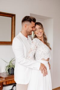 Ein Paar umarmt sich freudig in einem hellen Raum während seiner Hochzeit. Der Mann im weißen Anzug steht hinter der Frau, die ein weißes Spitzenkleid trägt. Beide lächeln und wirken glücklich. Im Hintergrund stehen ein Holztisch mit einer Pflanze und ein gerahmter Spiegel.