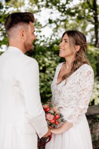Ein Paar steht draußen und lächelt sich in einer ruhigen, romantischen Umgebung an. Die Frau in einem weißen Spitzenkleid hält einen Blumenstrauß, während der Mann einen eleganten weißen Anzug trägt. Üppiges Grün umgibt sie und bildet die perfekte Kulisse für ihre Hochzeit.