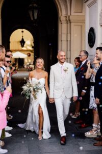 Eine Braut im weißen Kleid und ein Bräutigam im hellen Anzug gehen Hand in Hand und lächeln breit. Sie sind von einer fröhlichen Menge umgeben, die Seifenblasen oder Konfetti wirft, und feiern ihre Hochzeit vor einem reich verzierten Gebäudeeingang.