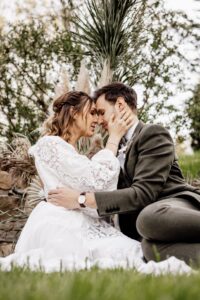 Ein Paar sitzt in romantischer Umarmung im Gras, ihre Stirnen berühren sich sanft. Die Frau im weißen Kleid hält das Gesicht des Mannes, umgeben von Grün und Pampasgras, und verkörpert den heiteren und liebevollen Geist einer Hochzeit.