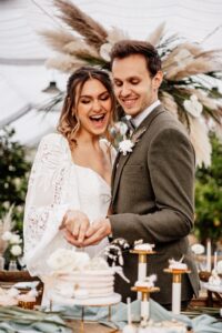 Ein glückliches Paar, das ganz in seine Hochzeit vertieft ist, lächelt, während es gemeinsam einen kleinen Kuchen anschneidet. Die Braut glänzt in einem weißen Spitzenkleid und der Bräutigam trägt einen grünen Anzug. Umgeben von elegantem Dekor, darunter Blumen und Pampasgras, war ihre Hochzeit perfekt und mit viel Flair geplant.