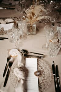 Elegante Tischdekoration für eine Hochzeit mit Kristallgläsern, einem transparenten Teller, Besteck und einer zu einem Knoten gebundenen Serviette. Unter dem Teller liegt ein Menü. In der Mitte verleihen ein dekoriertes Plätzchen und eine Vase mit Trockenblumen Ihrer besonderen Hochzeitsfeier Charme.