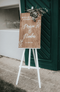 Ein Holzschild auf einer weißen Staffelei heißt die Gäste zur Hochzeit mit „Willkommen zu unserer Hochzeit“ und „Maria & Nicolas, 31. Juli 2021“ in weißer Schreibschrift willkommen. Dekorative Blätter zieren die Oberseite. Im Hintergrund verleihen eine grüne Tür und ein grünes Fenster diesem wunderschön geplanten Anlass Charme.