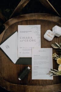 Ein Hochzeitseinladungsset für Chiara und Georg liegt auf einem Holzstuhl. Das Set enthält eine Einladungskarte, einen Umschlag und eine Karte mit Details. Daneben vervollständigen eine Spule mit grünem Garn, Blumendekor und eine kleine Schmuckschatulle mit Ringen das elegante Heiratsthema.