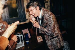 Eine Person in einer Jacke mit Leopardenmuster, die in einem schwach beleuchteten Innenraum lacht und in ein Retro-Telefon telefoniert, feiert ganz offensichtlich die Freuden einer Hochzeit. Eine andere Person ist teilweise zu sehen und gestikuliert lächelnd in ihre Richtung, was zur fröhlichen Atmosphäre dieses Hochzeitsereignisses beiträgt.