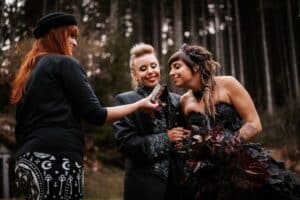 In einer unvergesslich schönen Waldkulisse tauscht ein Paar in gotischer Kleidung – einer in einem schwarzen Anzug und die andere in einem fließenden schwarzen Kleid – die Ringe aus. Eine dritte Person, gekleidet in rätselhafte Muster, assistiert bei der Hochzeitszeremonie. Im Hintergrund ragen majestätisch dunkle Bäume auf.
