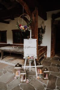 Ein rustikales Hochzeits-Willkommensschild auf einer mit Blumenarrangements geschmückten Staffelei gibt den Ton für Ihre Hochzeit an. Es ist von Laternen mit Kerzen umgeben, die ein gemütliches Licht verbreiten. Im Hintergrund sind eine Holzbank und eine rustikale Gebäudefassade zu sehen, perfekt für Ihre sorgfältig ausgearbeitete Hochzeitsplanung.
