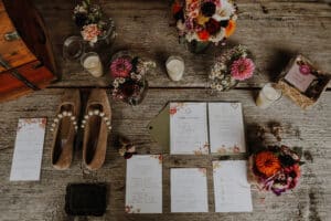 Ein rustikaler Holztisch ist mit Hochzeitseinladungen, Menüs und Antwortkarten mit Blumenmotiven geschmückt. Daneben runden braune Schuhe mit Perlendetails, Kerzen, Blumen in Töpfen und eine kleine braune Tasche die charmante Szene der Hochzeitsplanung ab.