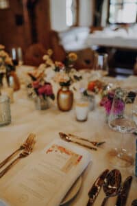 Ein wunderschön gedeckter Esstisch mit weißer Tischdecke, zusammengestellt für eine traumhafte Hochzeit. Er verfügt über ein Menü, goldenes Besteck, Weingläser und farbenfrohe Blumenarrangements in kleinen Vasen. Im sanft beleuchteten Hintergrund warten weitere Tische auf die Gäste als Teil einer sorgfältigen Hochzeitsplanung.