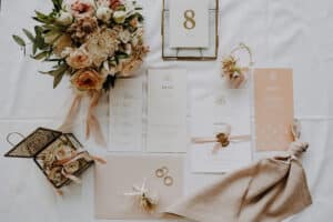 Eine flache Lage Hochzeitspapeterie auf einer weißen Oberfläche, die ein wunderschön zusammengestelltes Hochzeitsset präsentiert: Menü, Tischkarten und eine Einladung. Daneben liegen zwei goldene Ringe, ein Blumenstrauß, eine beige Serviette und eine dekorative Schachtel mit Blumen. Eine Tischnummernkarte mit „8“ vervollständigt die elegante Hochzeitsplanungspräsentation.