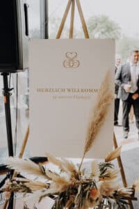 Auf einem Hochzeits-Willkommensschild mit goldener Schrift steht „Herzlich Willkommen zu unserer Hochzeit“. Es ist mit Pampasgras und Blumen geschmückt und spiegelt die Essenz einer durchdachten Hochzeitsplanung wider. Im Hintergrund sind ein Redner und Personen zu sehen, die die Szene für eine freudige Hochzeitsfeier vorbereiten.