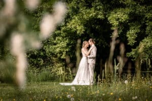 Ein Paar im Hochzeitskleid steht auf einer üppigen, grünen Wiese, umgeben von Wildblumen und dichten Bäumen. Sie umarmen sich unter sanftem, natürlichem Licht und schaffen so eine romantische Atmosphäre, die perfekt für ihre Hochzeit ist.