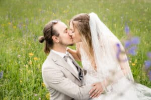 Braut und Bräutigam küssen sich auf einer üppigen grünen Wiese voller Wildblumen und fangen damit die Essenz ihrer Hochzeit ein. Die Braut trägt ein weißes Kleid mit Schleier und der Bräutigam einen beigen Anzug mit Sonnenbrille auf dem Kopf, während das Sonnenlicht diese romantische Szene sanft erhellt.