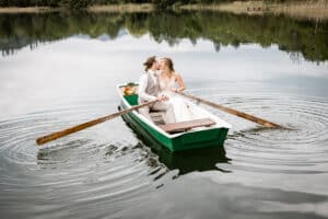 Ein Paar im Hochzeitskleid küsst sich, während es in einem kleinen grünen Ruderboot auf einem ruhigen See sitzt und verkörpert damit die Essenz von „Heiraten“. Die Braut hält einen Blumenstrauß, während sich im Wasser der bewölkte Himmel und die umliegenden Bäume spiegeln, und schafft so einen unvergesslichen Moment auf ihrer Hochzeitsreise.