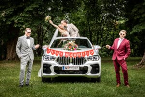 Ein frisch vermähltes Paar posiert verspielt auf einem weißen Auto, das mit einem „Just Married“-Banner geschmückt ist und die Freude ihrer Hochzeit einfängt. Die Braut hält einen Blumenstrauß, während zwei Männer in Anzügen, einer in Grau und einer in Rot, an ihrer Seite stehen. Die bezaubernde Szene spielt sich im Freien mit üppigem Grün als Hintergrund ab.