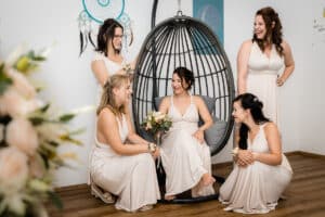 Fünf Frauen in fließenden, weißen Kleidern lächeln und sitzen um eine Frau herum, die in einem Hängesessel sitzt und die Essenz der Hochzeitsplanung verkörpert. Sie wirken entspannt und fröhlich und halten Blumensträuße in den Händen. Im Hintergrund sind ein Traumfänger und zarte Blumendekorationen zu sehen, perfekt für einen herzlichen Hochzeitsmoment.