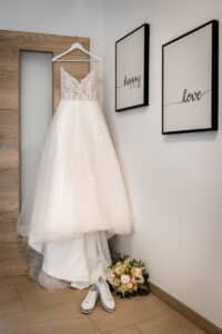 An einer Holztür hängt ein weißes Brautkleid, das den Beginn einer umfassenden Hochzeitsplanung symbolisiert. Darunter stehen weiße Turnschuhe neben einem Blumenstrauß. Auf zwei gerahmten Drucken an der Wand stehen „Happy“ und „Love“.